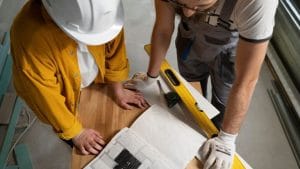 Dos personas con casco y guantes revisan planos de construcción sobre una mesa de madera, junto a herramientas como un nivel y una cinta métrica.